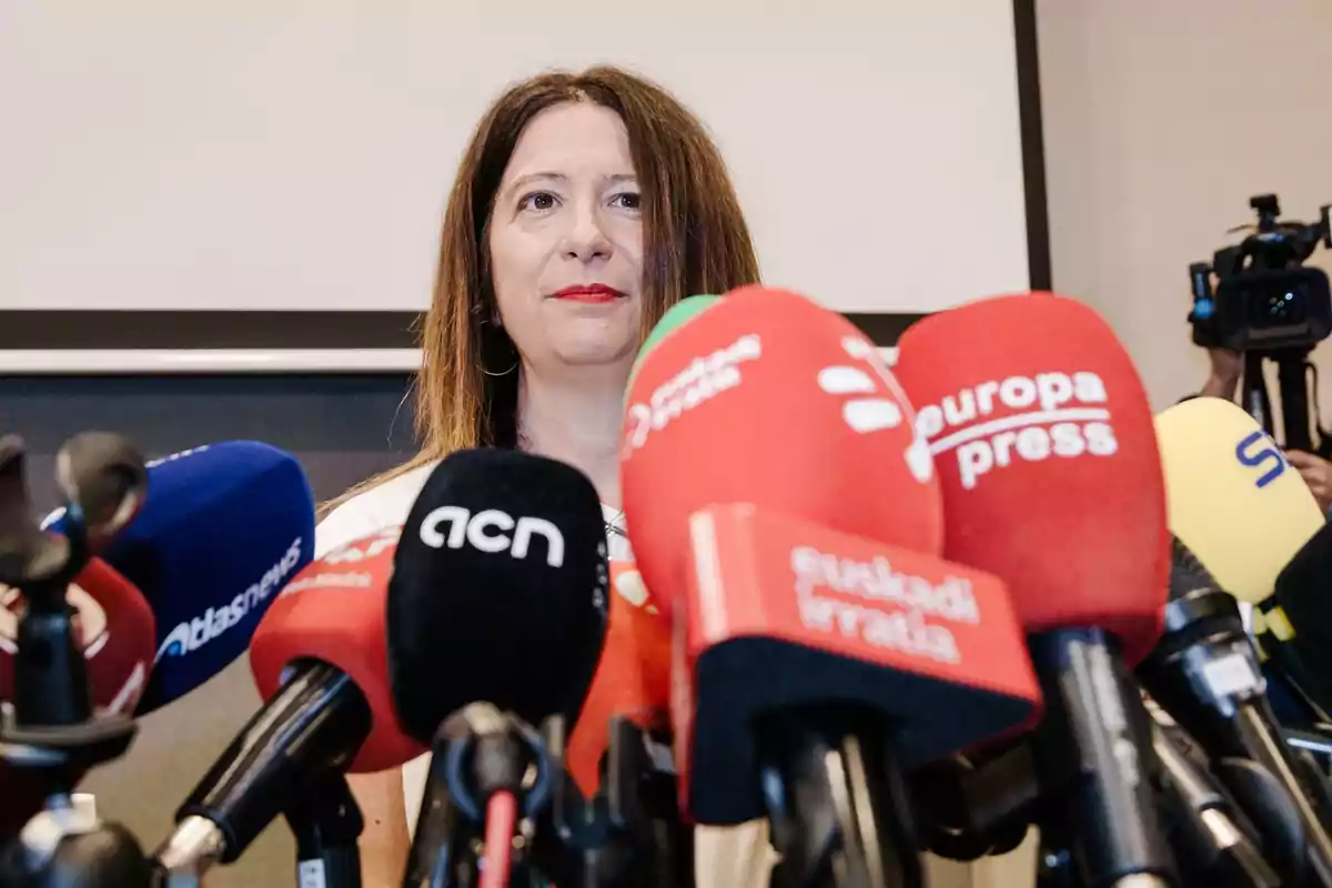Mujer de cabello largo y lacio hablando frente a varios micrófonos de diferentes medios de comunicación durante una rueda de prensa Mujer de cabello largo y lacio hablando frente a varios micrófonos de diferentes medios de comunicación durante una rueda de prensa
