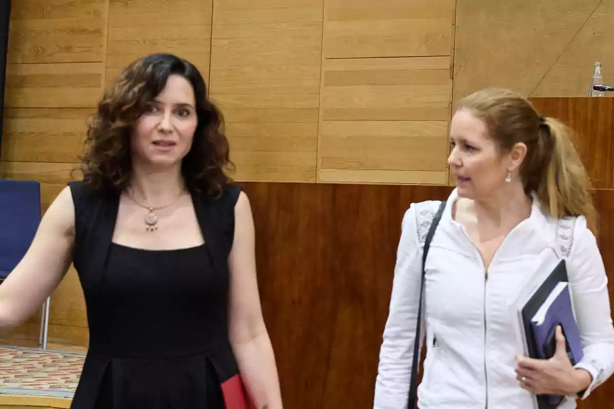 Dos mujeres conversan en un interior con paredes de madera, una lleva vestido negro y la otra chaqueta blanca y sostiene carpetas.