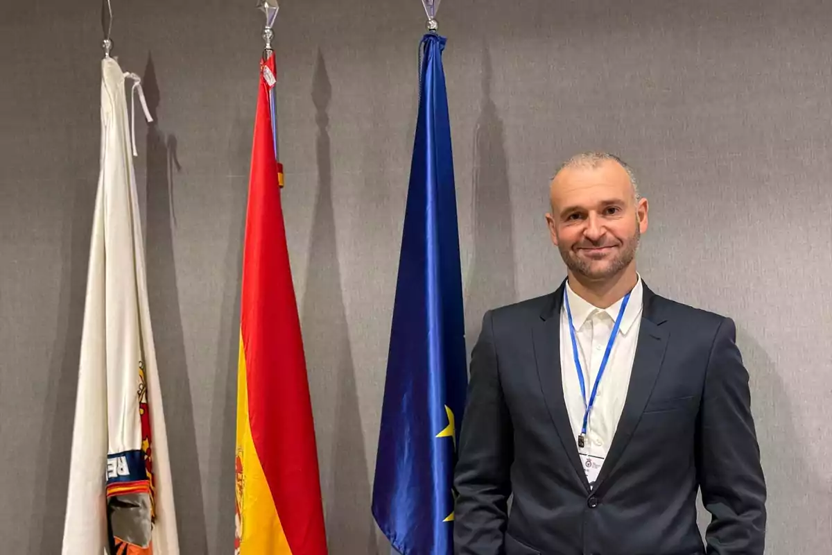 Hombre con traje oscuro y gafete posando junto a tres banderas en un entorno formal