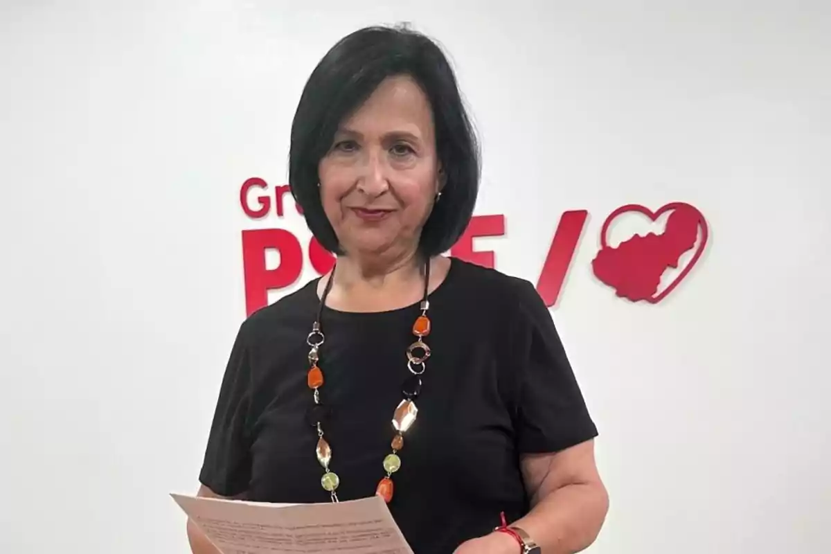 Mujer de cabello corto y oscuro con camiseta negra y collar de cuentas de colores sosteniendo unos papeles frente a una pared blanca con el logo del PSOE y un corazón rojo Mujer de cabello corto y oscuro con camiseta negra y collar de cuentas de colores sosteniendo unos papeles frente a una pared blanca con el logo del PSOE y un corazón rojo