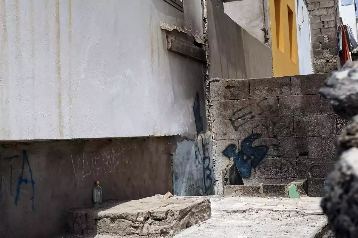 Paredes de concreto con grafitis y manchas, una botella de vidrio en el suelo y una casa con ventana amarilla al fondo