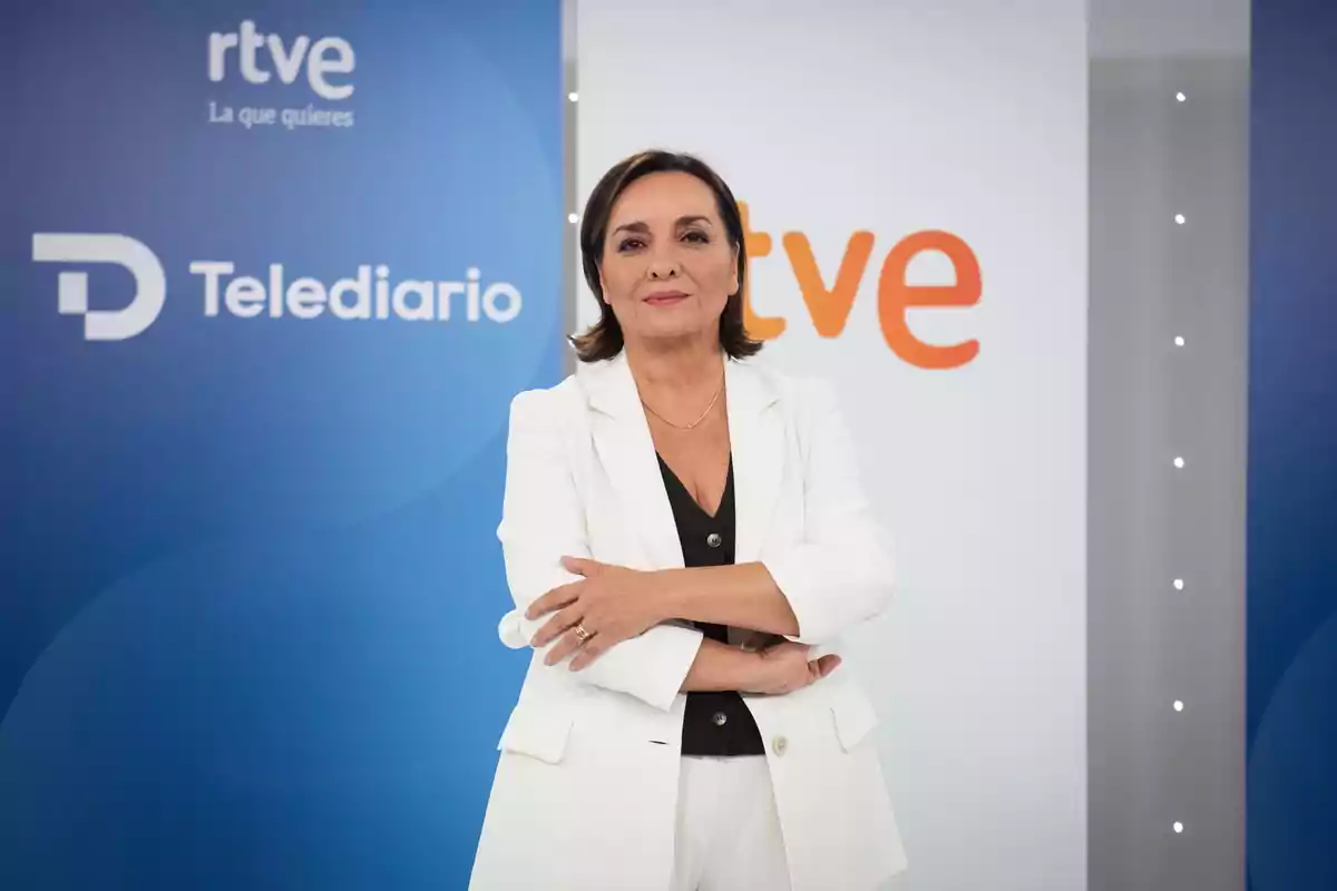 Mujer de cabello corto y oscuro vestida con traje blanco posando con los brazos cruzados frente a un fondo con los logotipos de Telediario y rtve
