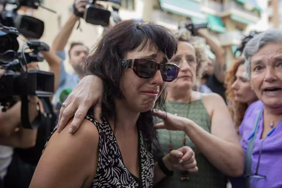 Mujer con gafas de sol llorando y siendo consolada por otras personas mientras varias cámaras la rodean en un ambiente de tensión y emoción