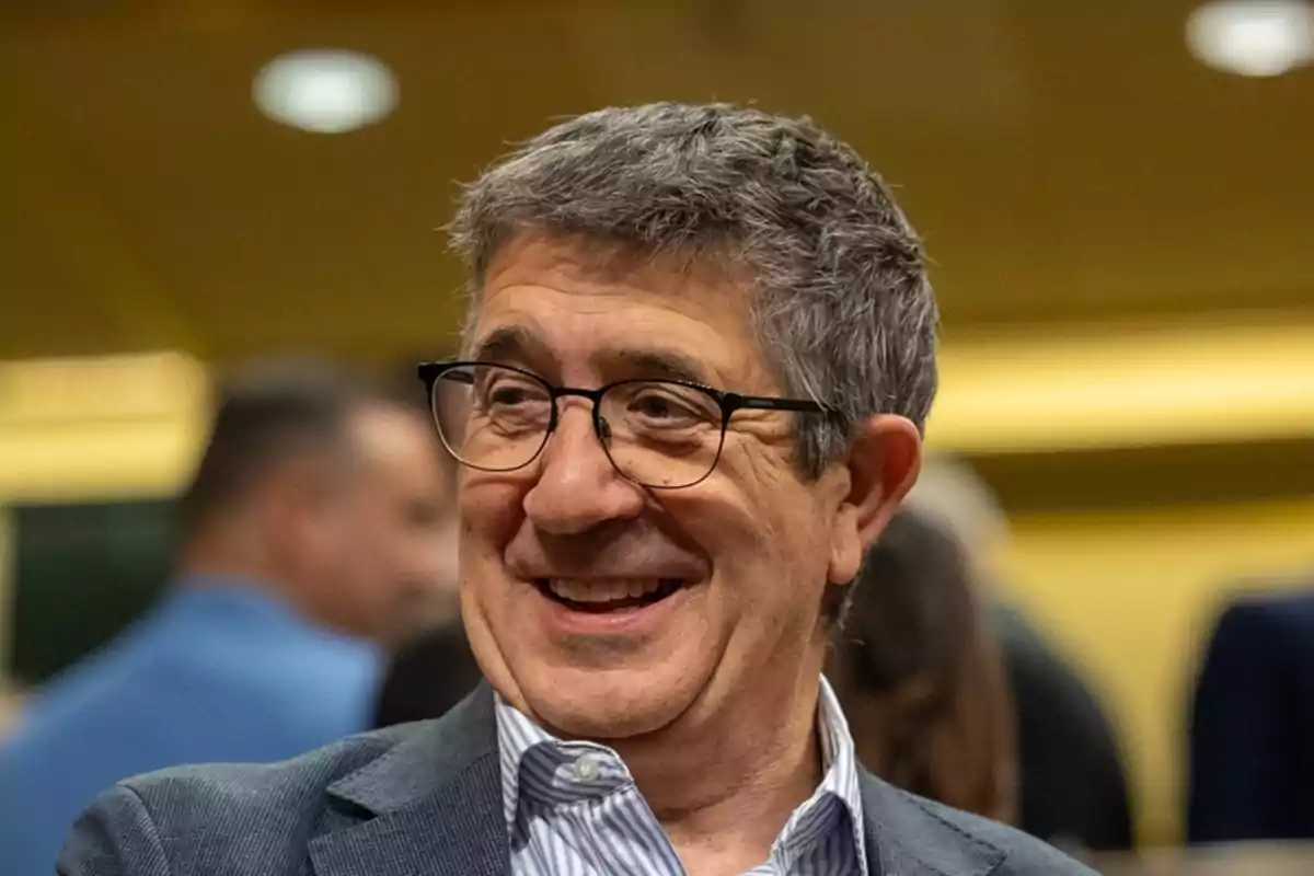 Hombre de cabello canoso y gafas sonriendo en un ambiente interior con personas al fondo desenfocadas Hombre de cabello canoso y gafas sonriendo en un ambiente interior con personas al fondo desenfocadas