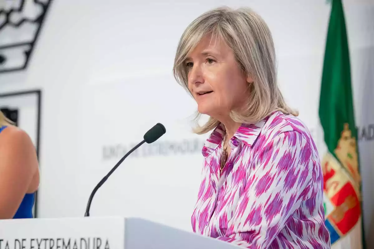 Mujer de cabello rubio y camisa estampada en tonos rosas hablando frente a un micrófono en un atril con una bandera verde al fondo Mujer de cabello rubio y camisa estampada en tonos rosas hablando frente a un micrófono en un atril con una bandera verde al fondo