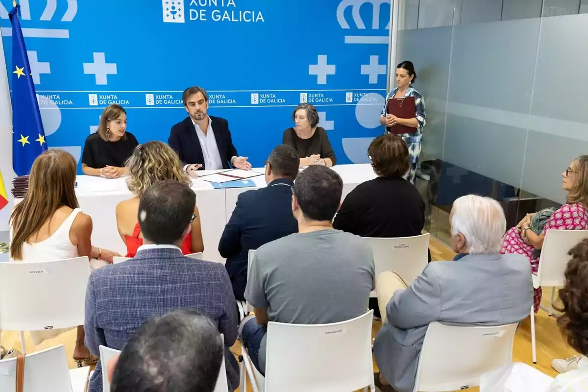 Varias personas asisten a una reunión en una sala con fondo azul de la Xunta de Galicia y banderas oficiales, mientras tres personas presiden la mesa principal y una mujer de pie sostiene una carpeta. Varias personas asisten a una reunión en una sala con fondo azul de la Xunta de Galicia y banderas oficiales, mientras tres personas presiden la mesa principal y una mujer de pie sostiene una carpeta.