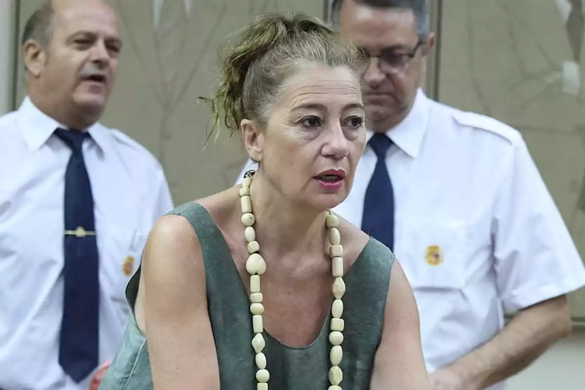 Mujer con collar de cuentas grandes y vestido verde, acompañada por dos hombres con camisa blanca y corbata azul en un ambiente interior Mujer con collar de cuentas grandes y vestido verde, acompañada por dos hombres con camisa blanca y corbata azul en un ambiente interior