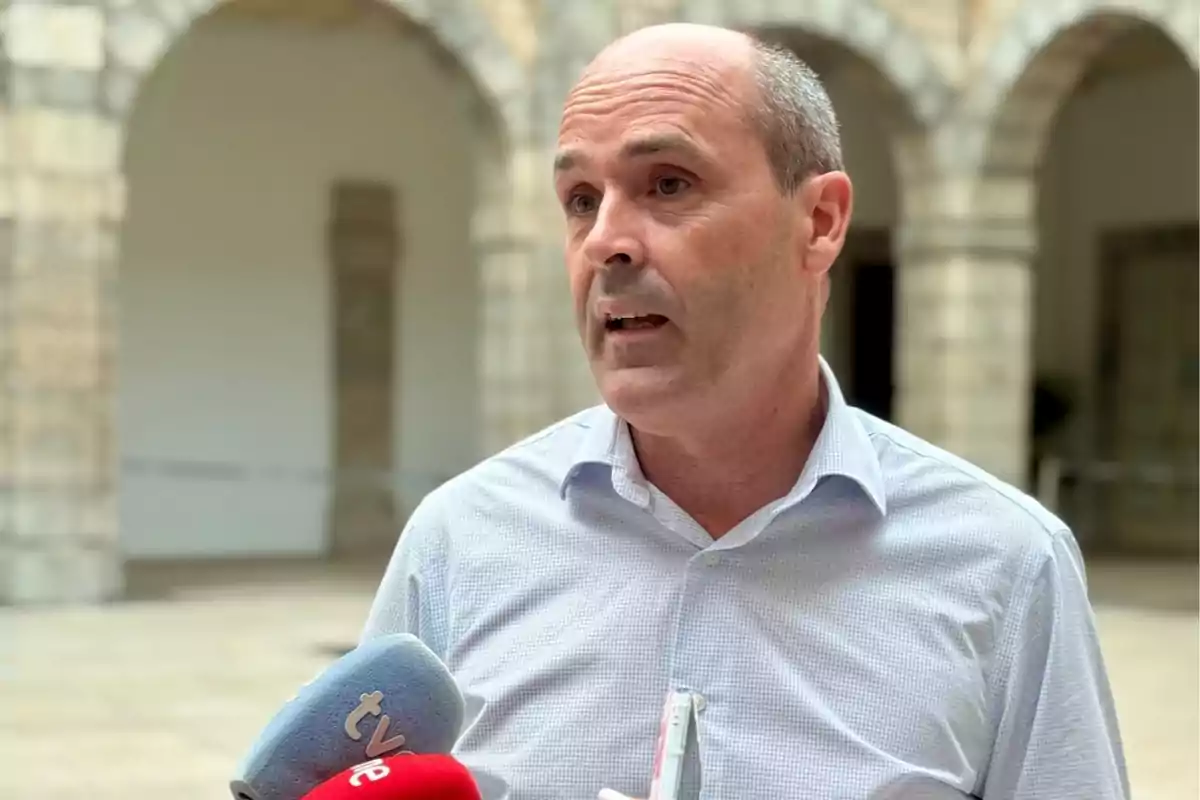Hombre calvo con camisa clara hablando frente a varios micrófonos en un patio con arcos de piedra al fondo Hombre calvo con camisa clara hablando frente a varios micrófonos en un patio con arcos de piedra al fondo