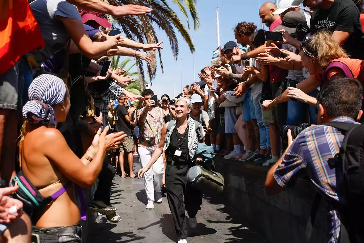 Una multitud aplaude y recibe con entusiasmo a una persona sonriente que camina entre ellos bajo el sol, rodeada de palmeras y ambiente festivo. Una multitud aplaude y recibe con entusiasmo a una persona sonriente que camina entre ellos bajo el sol, rodeada de palmeras y ambiente festivo.