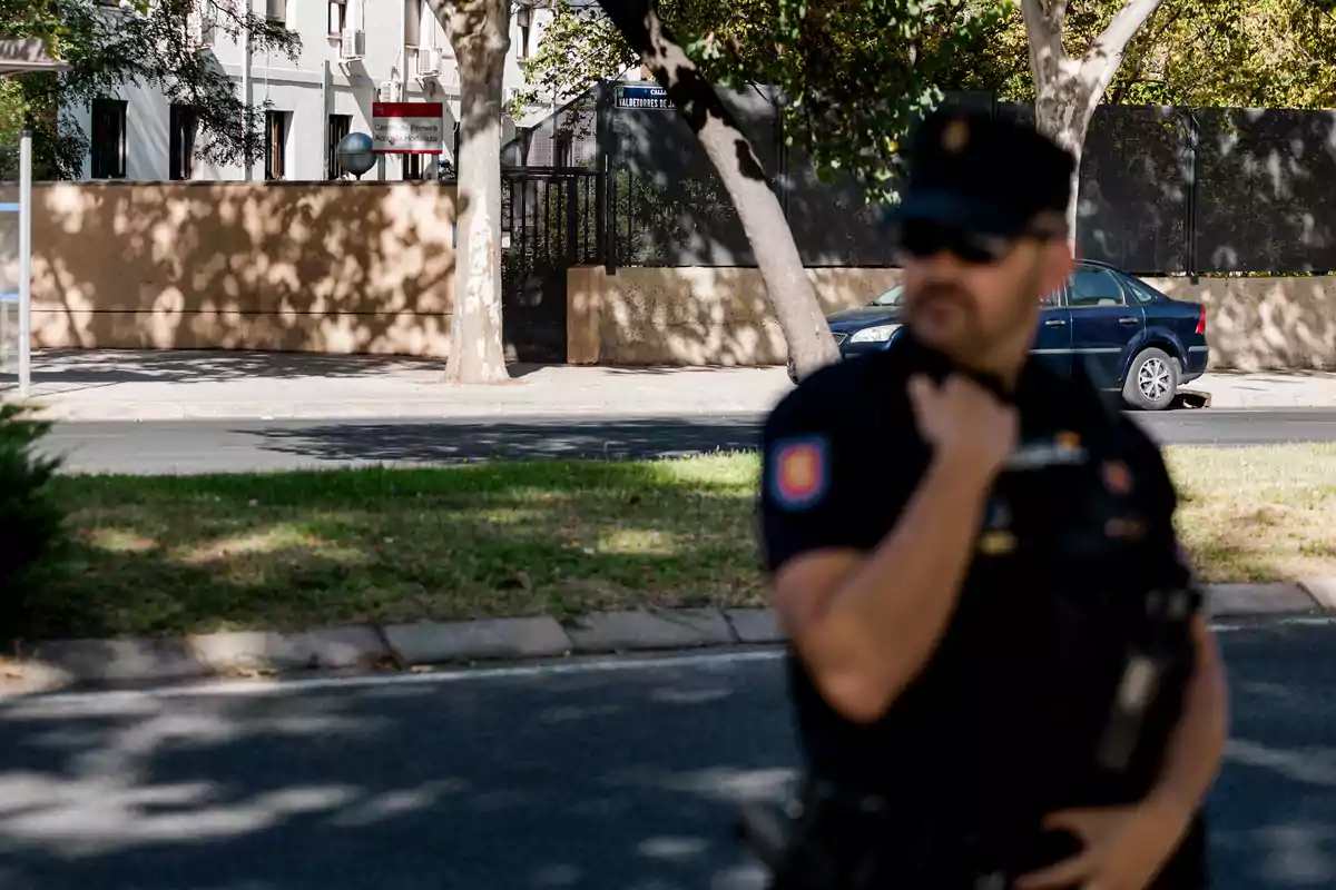 Policía de pie en primer plano con uniforme oscuro y gafas de sol mientras al fondo se observa un edificio, árboles y un coche azul estacionado Policía de pie en primer plano con uniforme oscuro y gafas de sol mientras al fondo se observa un edificio, árboles y un coche azul estacionado