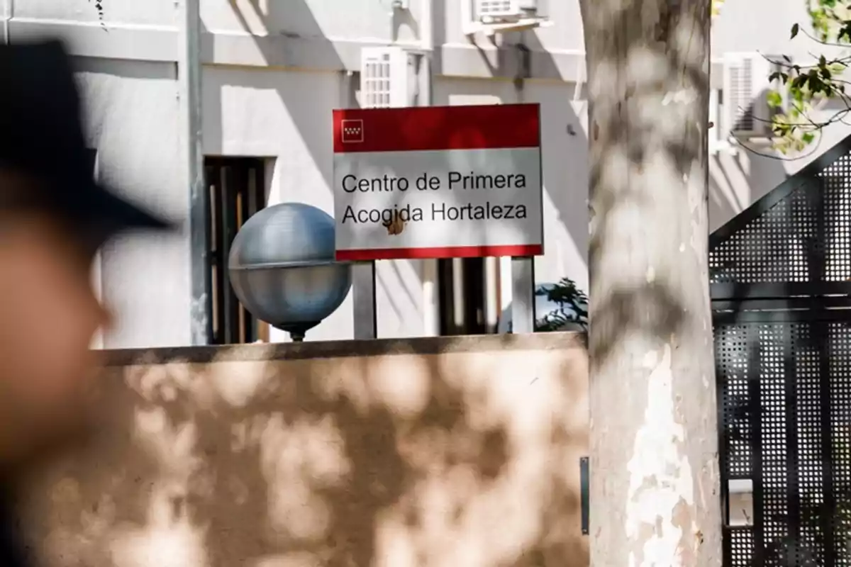 Cartel del Centro de Primera Acogida Hortaleza en la entrada de un edificio con una persona desenfocada en primer plano y un árbol al lado Cartel del Centro de Primera Acogida Hortaleza en la entrada de un edificio con una persona desenfocada en primer plano y un árbol al lado