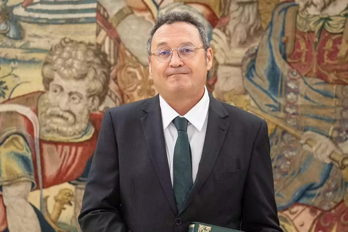 Hombre de traje oscuro y corbata verde posando frente a un tapiz decorativo con figuras históricas