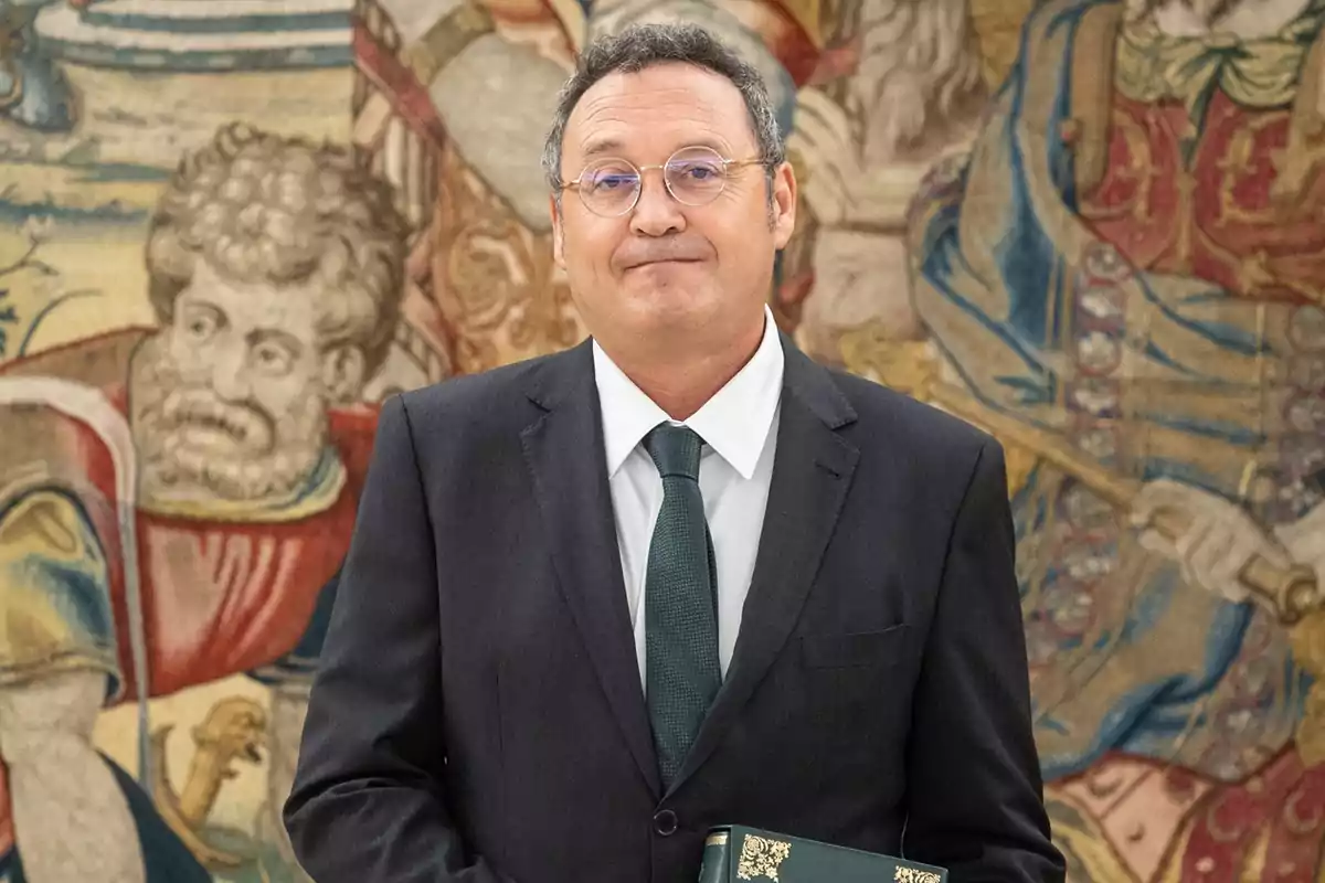 Hombre con gafas y traje oscuro sosteniendo un libro frente a un tapiz decorativo
