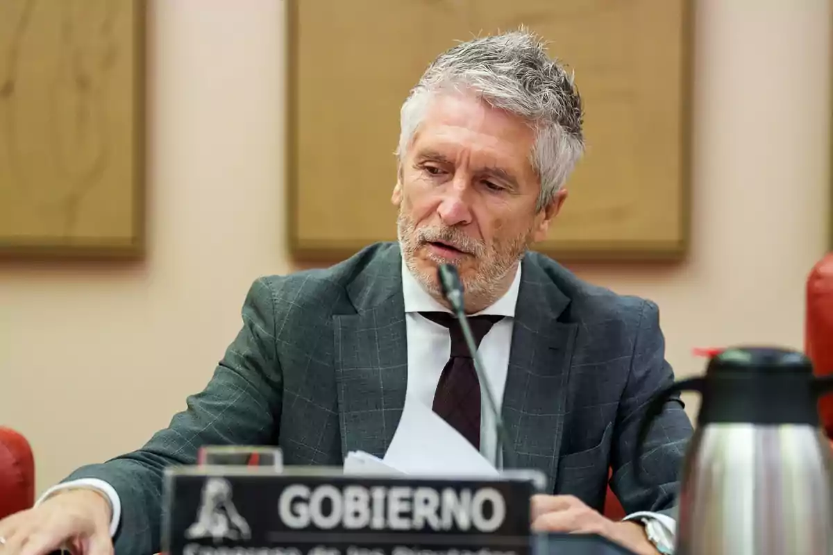 Hombre de cabello canoso y barba sentado en una mesa con un micrófono y un cartel que dice gobierno mientras revisa unos papeles