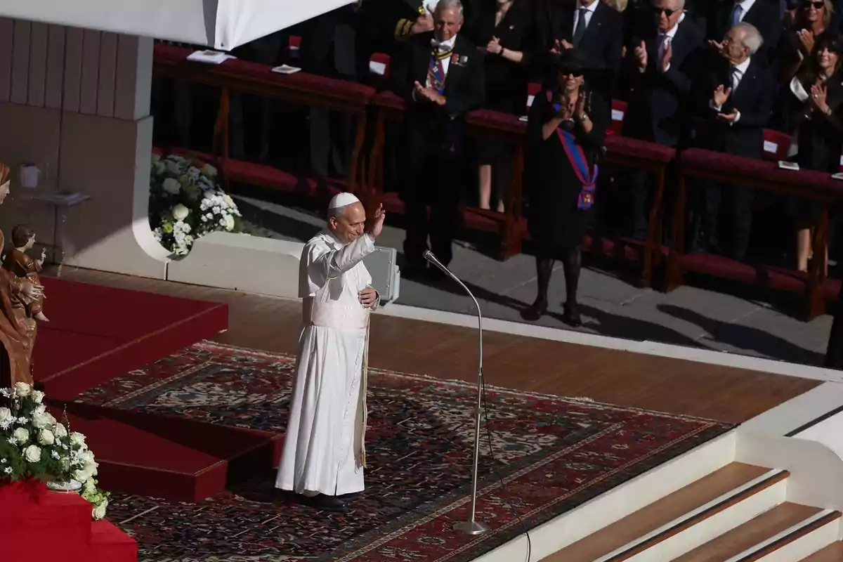 El Papa saluda a los asistentes desde un escenario decorado con alfombra y flores mientras varias personas aplauden de pie en el fondo El Papa saluda a los asistentes desde un escenario decorado con alfombra y flores mientras varias personas aplauden de pie en el fondo