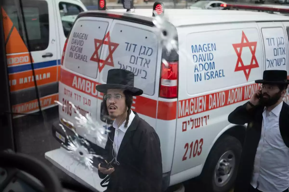 Dos hombres con vestimenta tradicional judía ortodoxa están junto a una ambulancia de Maguen David Adom en Israel y un vehículo de emergencia, mientras el parabrisas frente a ellos muestra impactos de bala. Dos hombres con vestimenta tradicional judía ortodoxa están junto a una ambulancia de Maguen David Adom en Israel y un vehículo de emergencia, mientras el parabrisas frente a ellos muestra impactos de bala.
