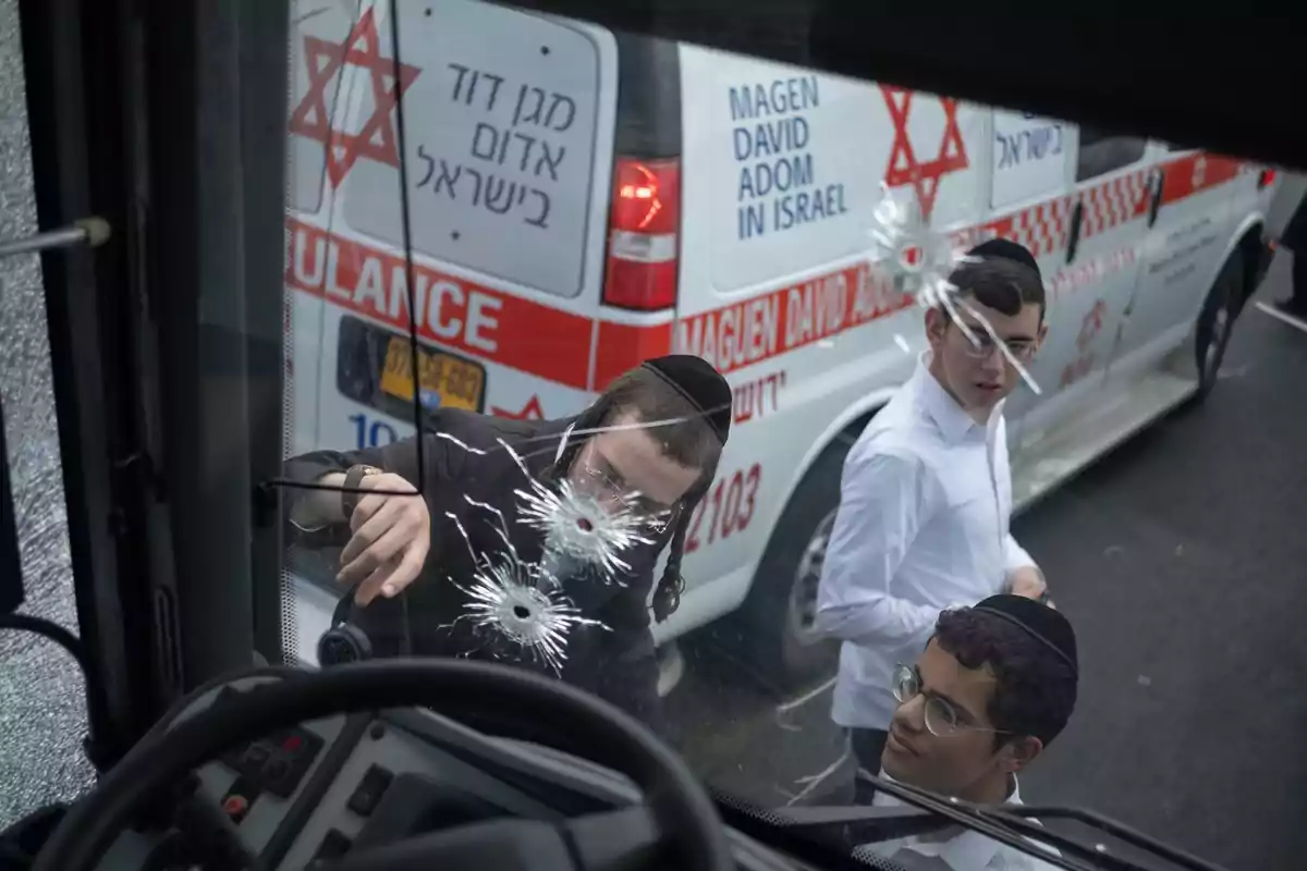 Tres jóvenes observan el parabrisas de un vehículo con impactos de bala mientras una ambulancia israelí está estacionada al fondo Tres jóvenes observan el parabrisas de un vehículo con impactos de bala mientras una ambulancia israelí está estacionada al fondo