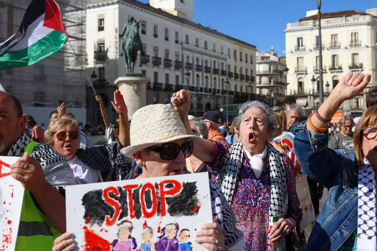 Personas mayores protestan en una plaza con pancartas y una bandera palestina mientras levantan el puño y muestran expresiones de indignación