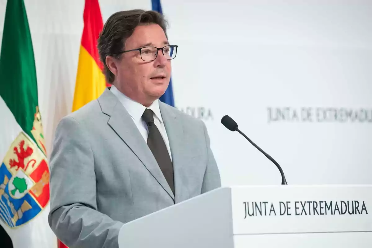 Hombre con gafas y traje gris hablando en un atril de la Junta de Extremadura con banderas al fondo
