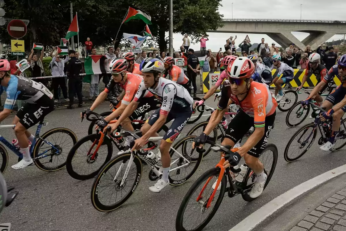 Ciclistas compitiendo en una carrera mientras un grupo de personas con banderas observa y anima desde el borde de la calle