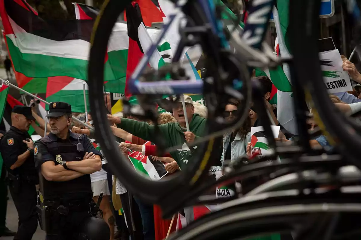 Manifestación con banderas palestinas y pancartas, un grupo de personas protesta mientras un policía observa y una bicicleta aparece en primer plano desenfocada