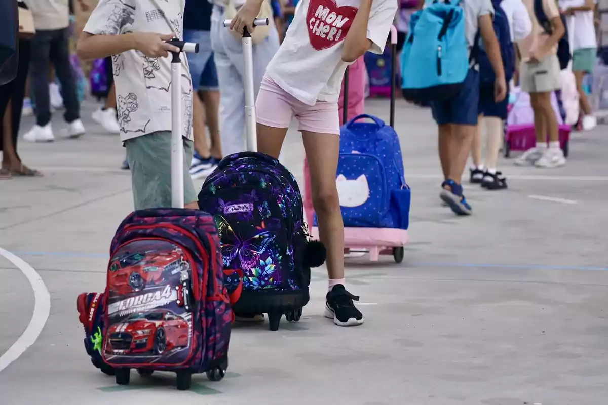 Niños con mochilas de ruedas en un patio escolar rodeados de otros estudiantes Niños con mochilas de ruedas en un patio escolar rodeados de otros estudiantes