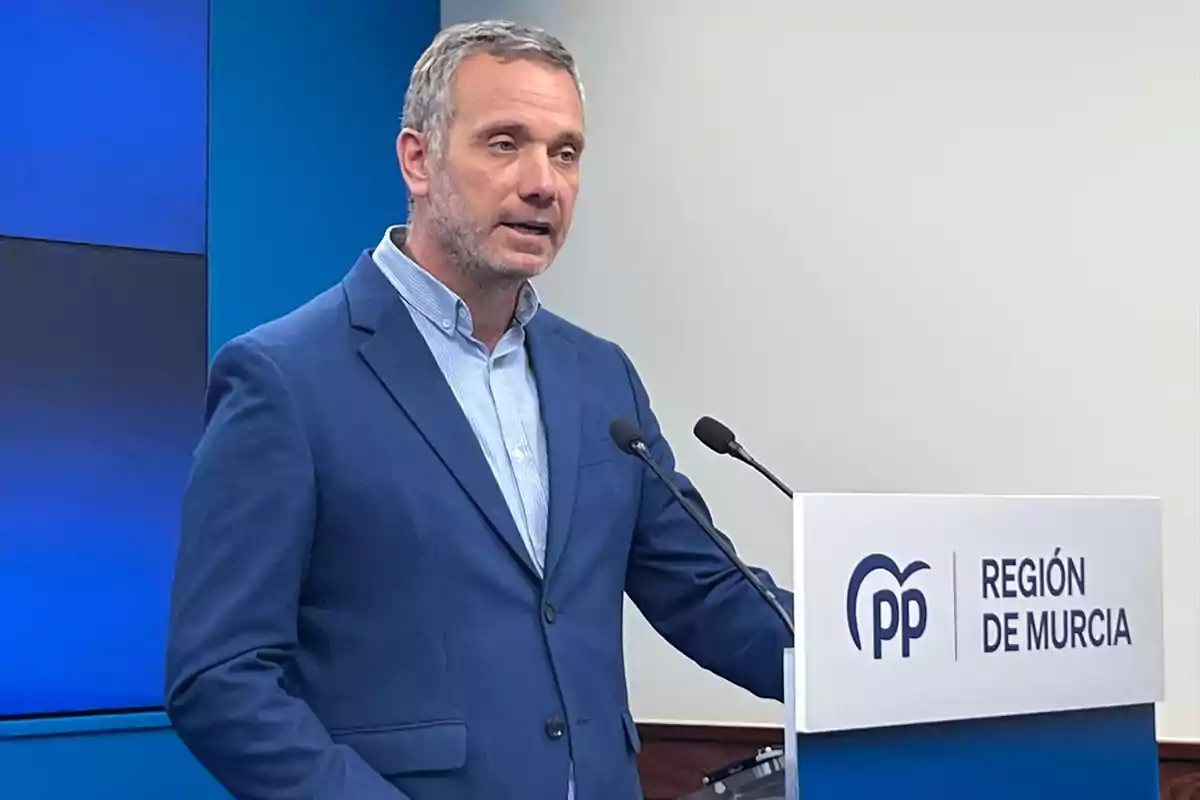 Hombre de cabello corto y canoso con barba hablando en un atril con el logo del PP y el texto Región de Murcia