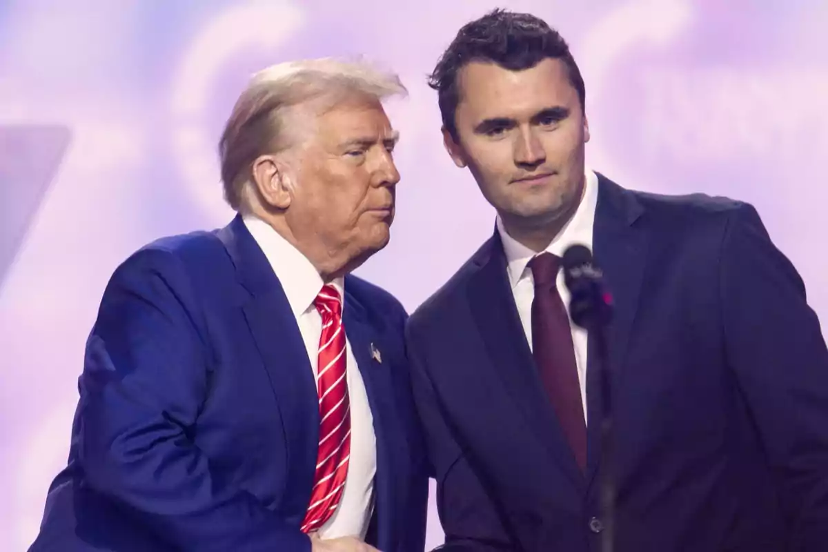 Dos hombres con traje conversan en un escenario con fondo morado y un micrófono al frente