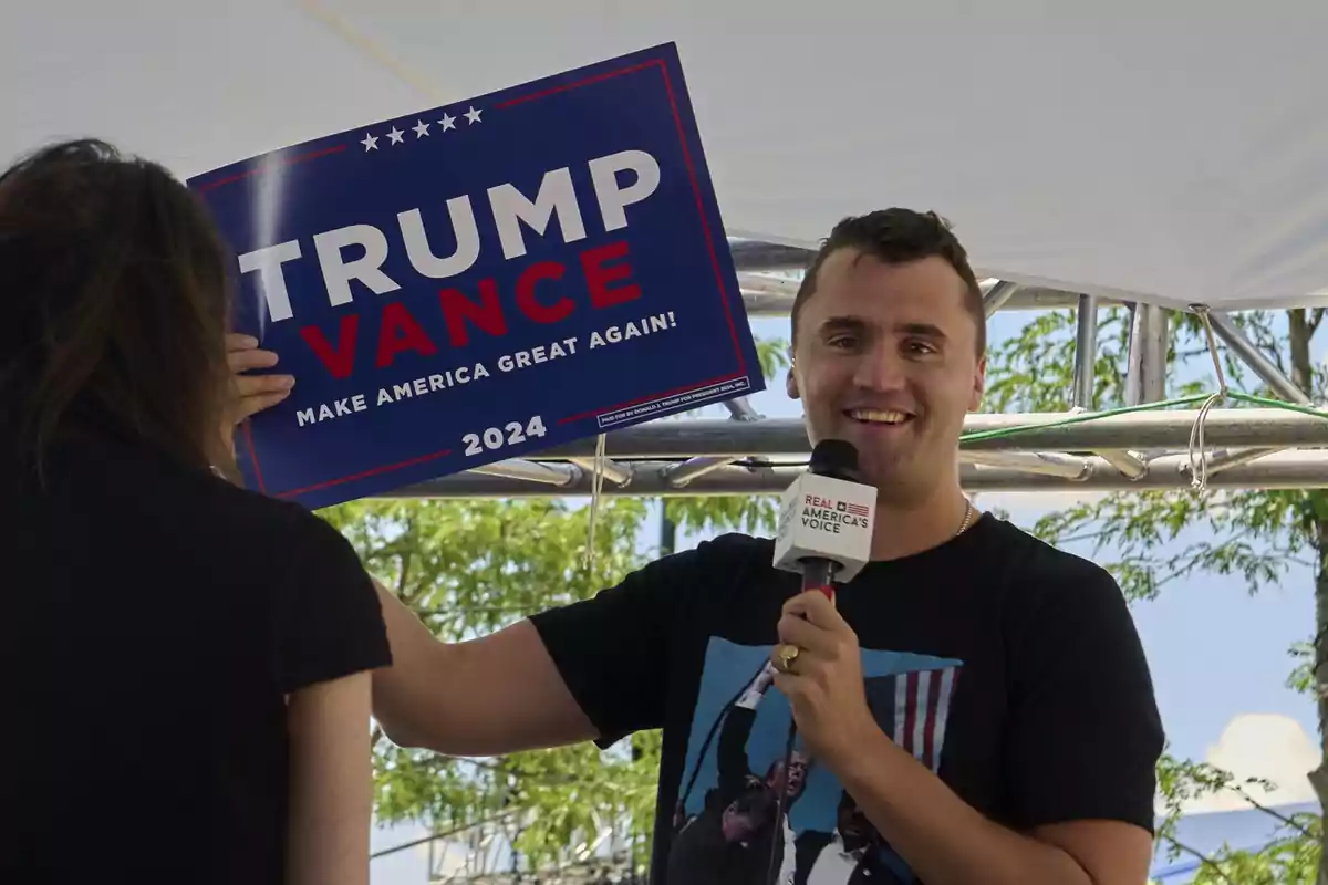 Hombre sonriente sostiene un micrófono y un cartel de campaña de Trump Vance 2024 junto a una mujer bajo una carpa al aire libre