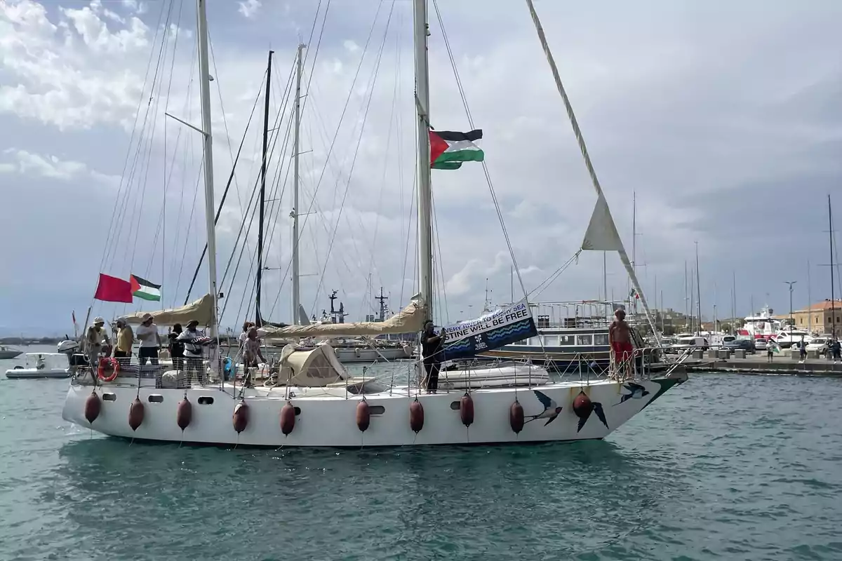 Un velero con varias personas a bordo navega cerca de un puerto, lleva banderas y una pancarta que apoya la libertad de Palestina.