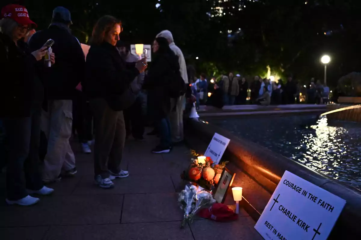 Personas reunidas de noche junto a una fuente, algunas sosteniendo velas, con flores, carteles y velas encendidas en el suelo en señal de homenaje.