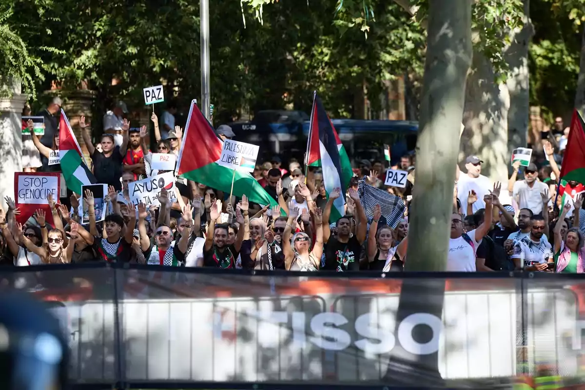 Manifestación con personas levantando las manos, ondeando banderas palestinas y sosteniendo carteles que piden paz y el fin del genocidio, en un entorno al aire libre con árboles al fondo