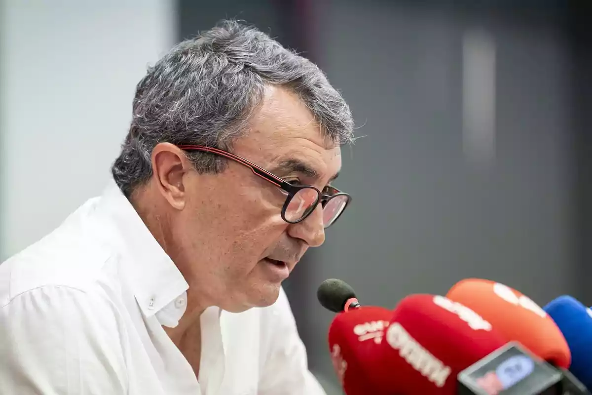 Hombre de cabello canoso y gafas hablando frente a varios micrófonos en una rueda de prensa