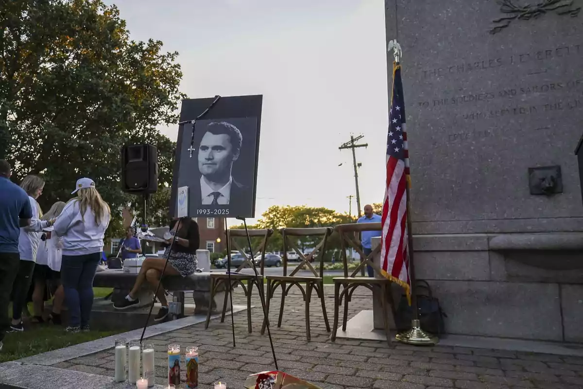 Personas reunidas en una vigilia al aire libre junto a un retrato en blanco y negro de un hombre con fechas, varias velas encendidas y una bandera de Estados Unidos junto a un monumento de piedra Personas reunidas en una vigilia al aire libre junto a un retrato en blanco y negro de un hombre con fechas, varias velas encendidas y una bandera de Estados Unidos junto a un monumento de piedra