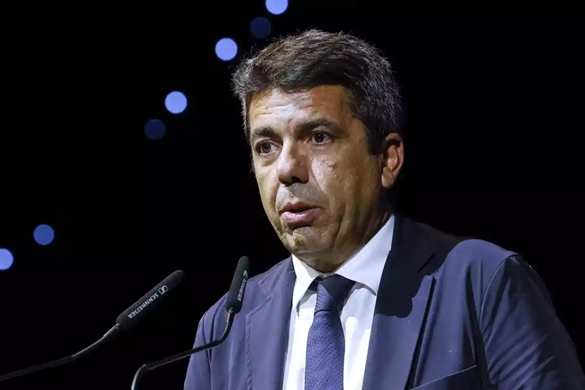 Hombre de cabello corto y canoso hablando frente a dos micrófonos con fondo oscuro y luces desenfocadas