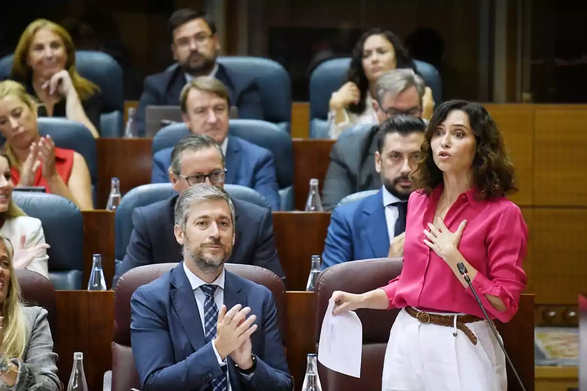 Una mujer de cabello rizado y camisa rosa habla de pie en una sala de reuniones mientras varias personas la observan y aplauden Una mujer de cabello rizado y camisa rosa habla de pie en una sala de reuniones mientras varias personas la observan y aplauden