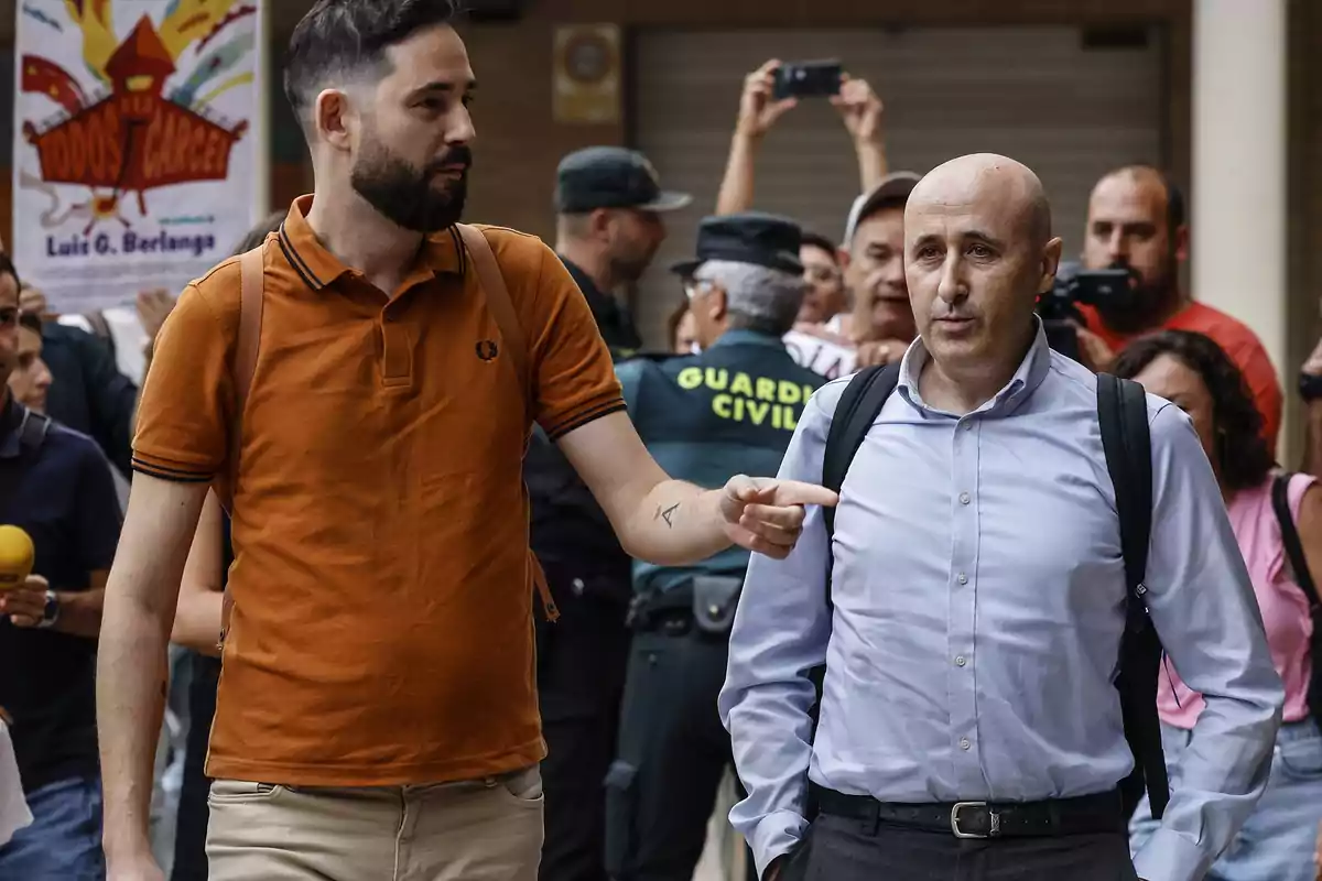 Un hombre con camisa azul y mochila camina junto a otro hombre con camiseta naranja mientras varias personas y agentes de la Guardia Civil están al fondo