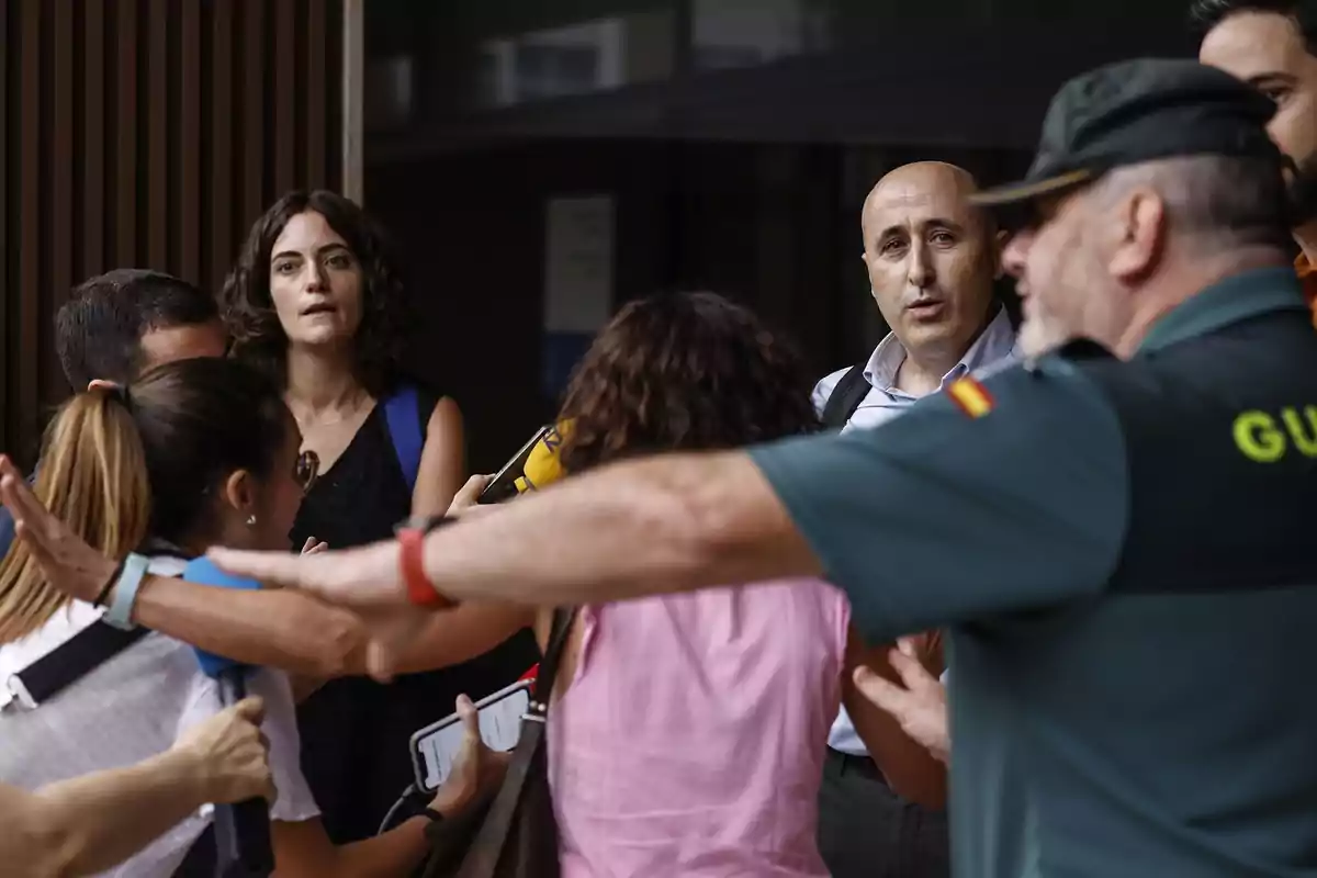 Un guardia civil intenta contener a un grupo de periodistas que rodean a dos personas a la entrada de un edificio