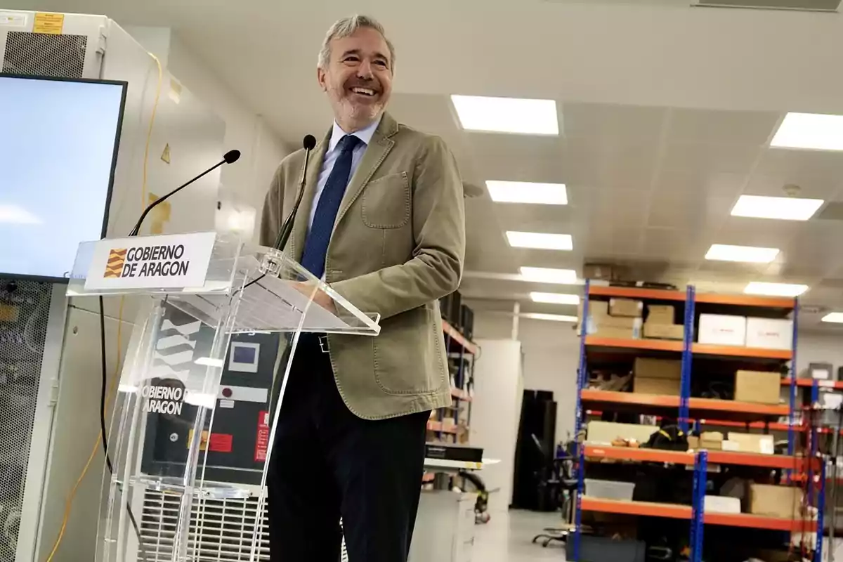 Hombre sonriente con chaqueta beige y corbata azul hablando en un atril del Gobierno de Aragón dentro de una sala con estanterías y cajas al fondo Hombre sonriente con chaqueta beige y corbata azul hablando en un atril del Gobierno de Aragón dentro de una sala con estanterías y cajas al fondo