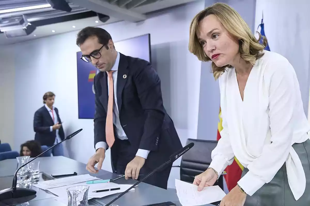 Dos personas en una sala de conferencias preparándose para una rueda de prensa con micrófonos y papeles sobre la mesa y una bandera de España al fondo