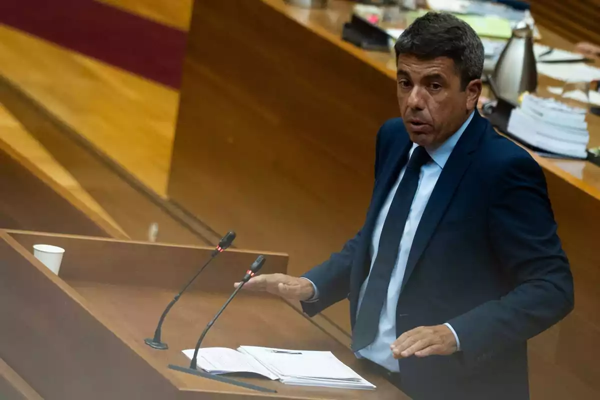 Hombre de traje azul hablando en un atril de madera con micrófonos y documentos en una sala de reuniones Hombre de traje azul hablando en un atril de madera con micrófonos y documentos en una sala de reuniones