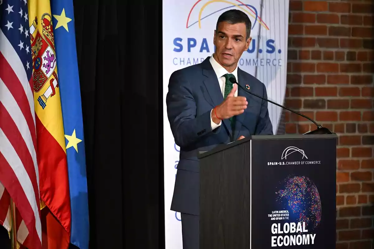 Hombre de traje hablando en un podio durante un evento de la Cámara de Comercio España-Estados Unidos con banderas de Estados Unidos, España y la Unión Europea al fondo Hombre de traje hablando en un podio durante un evento de la Cámara de Comercio España-Estados Unidos con banderas de Estados Unidos, España y la Unión Europea al fondo