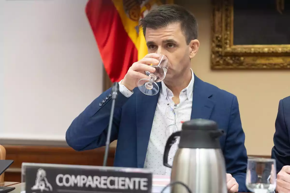 Hombre con chaqueta azul bebiendo agua en una reunión oficial con una bandera de España al fondo