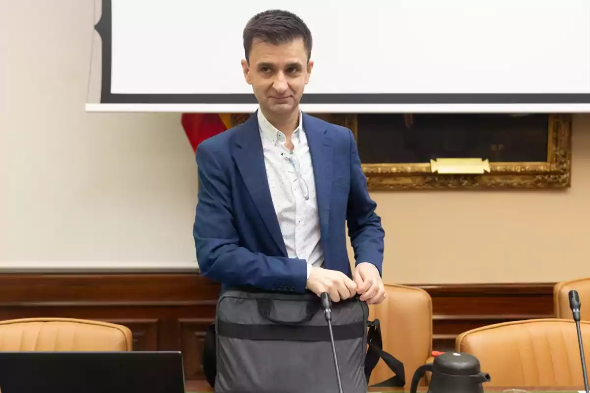 Hombre de pie en una sala de reuniones con saco azul y camisa clara, sosteniendo una bolsa de mano frente a una mesa con micrófono y computadora portátil