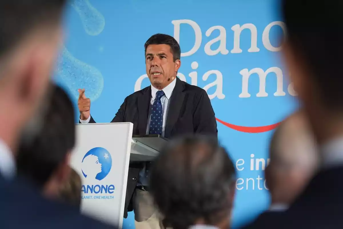 Hombre de traje hablando en un podio de Danone frente a una audiencia con fondo azul y letras blancas Hombre de traje hablando en un podio de Danone frente a una audiencia con fondo azul y letras blancas