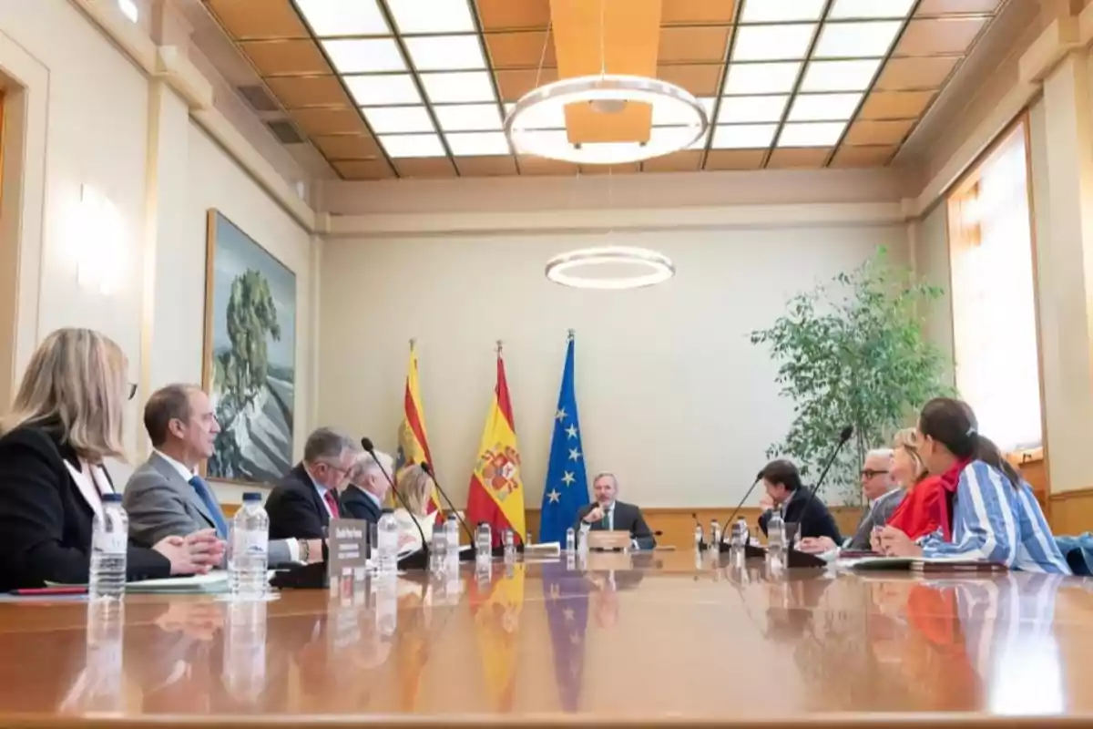 Varias personas sentadas alrededor de una mesa de reuniones en una sala oficial con banderas de España y la Unión Europea al fondo Varias personas sentadas alrededor de una mesa de reuniones en una sala oficial con banderas de España y la Unión Europea al fondo