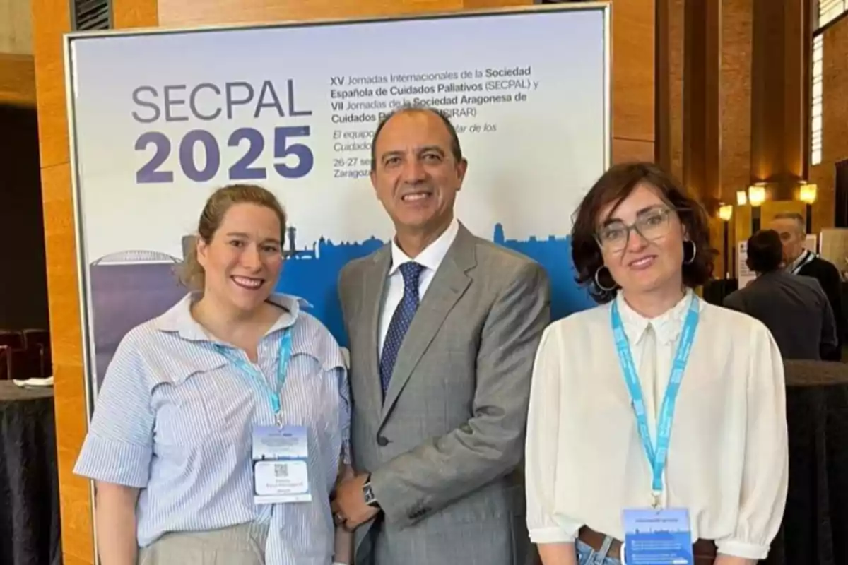 Tres personas posan sonrientes frente a un cartel de un congreso de cuidados paliativos llamado SECPAL 2025 en un ambiente de conferencia. Tres personas posan sonrientes frente a un cartel de un congreso de cuidados paliativos llamado SECPAL 2025 en un ambiente de conferencia.