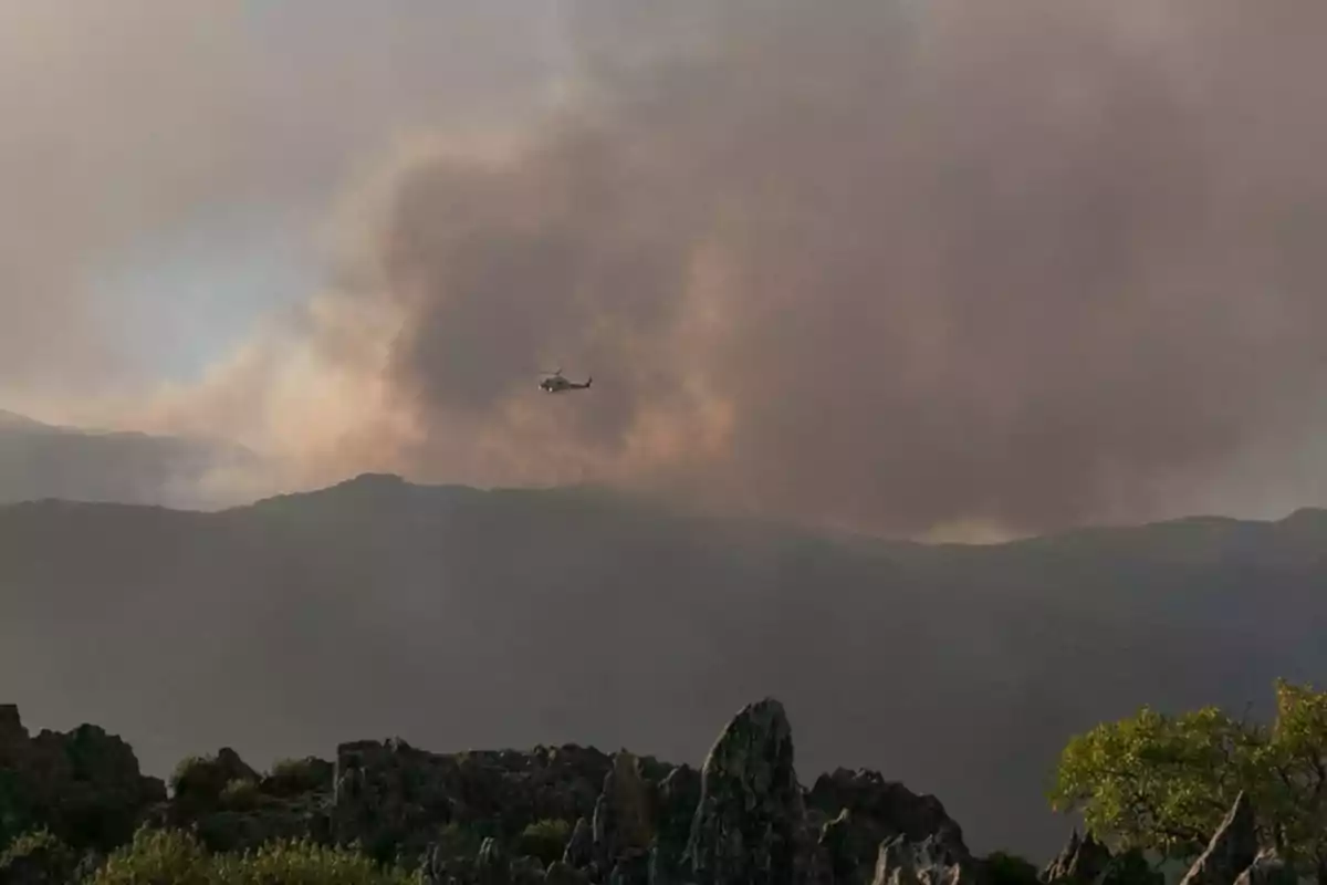 Helicóptero sobrevolando una zona montañosa con una gran nube de humo en el fondo