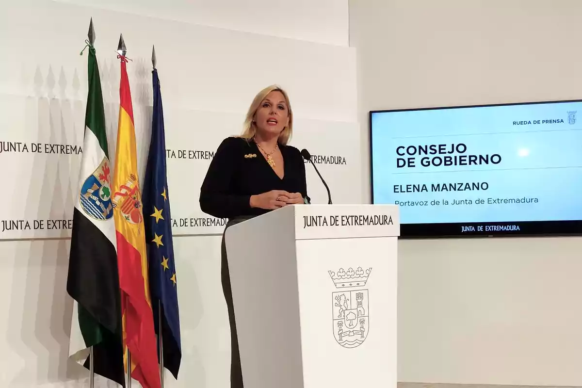 Una mujer rubia con chaqueta negra habla en un atril blanco con el escudo de la Junta de Extremadura, junto a tres banderas y una pantalla que muestra información sobre una rueda de prensa del Consejo de Gobierno Una mujer rubia con chaqueta negra habla en un atril blanco con el escudo de la Junta de Extremadura, junto a tres banderas y una pantalla que muestra información sobre una rueda de prensa del Consejo de Gobierno