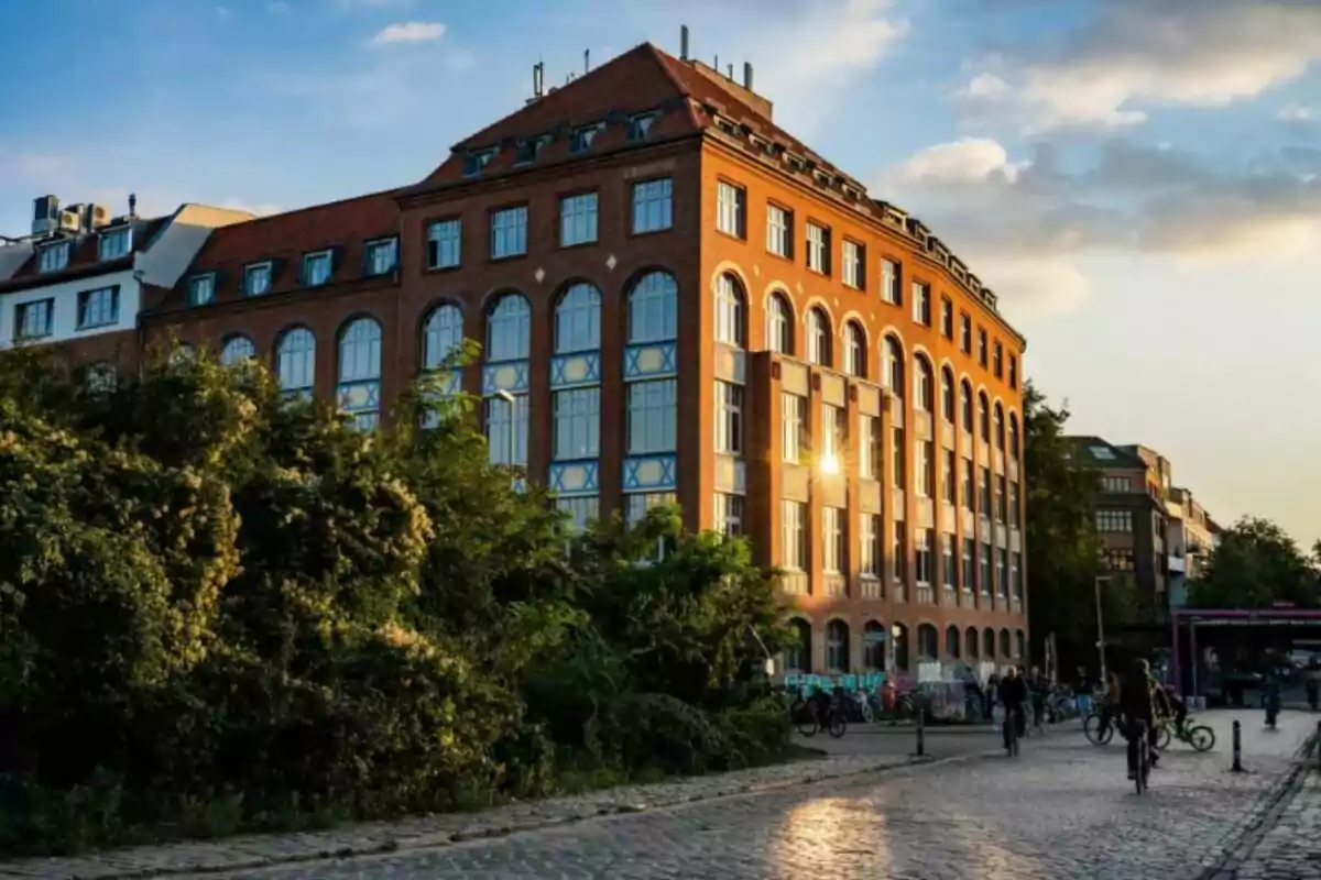 Edificio de ladrillo con grandes ventanas reflejando la luz del atardecer, rodeado de vegetación y personas andando en bicicleta por una calle empedrada Edificio de ladrillo con grandes ventanas reflejando la luz del atardecer, rodeado de vegetación y personas andando en bicicleta por una calle empedrada